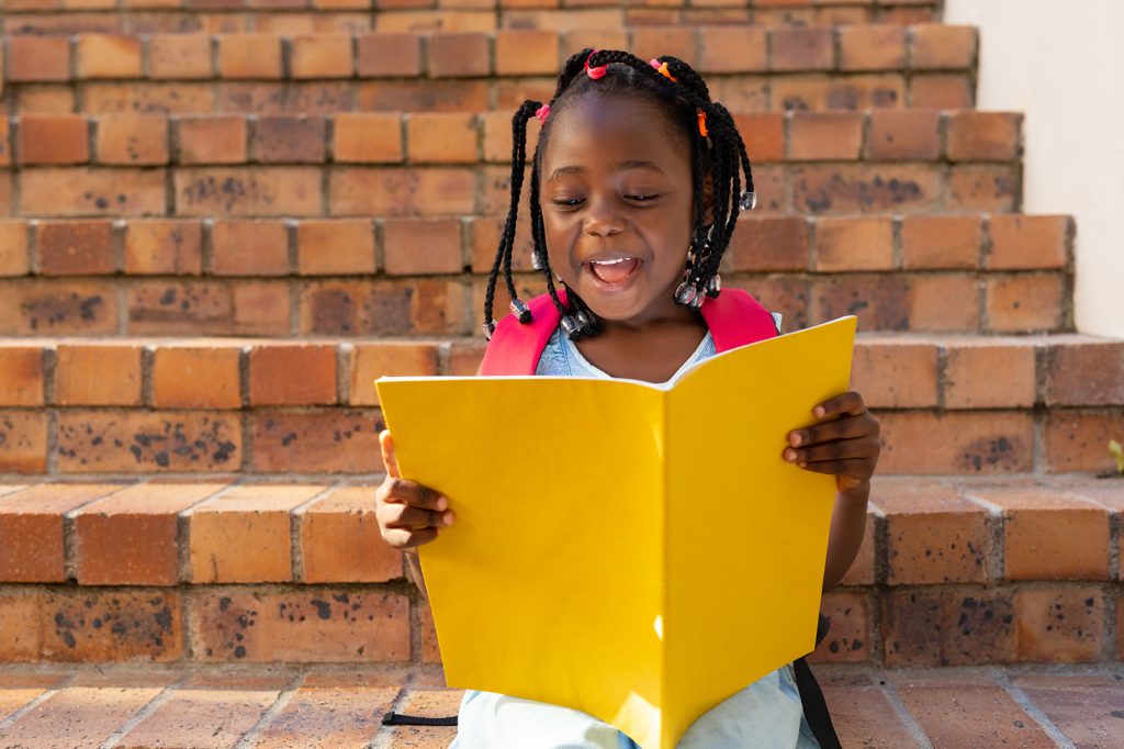 Happy african american schoolgirl sitting on stairs and reading book at elementary school. Education, childhood, development, learning and school, unaltered.